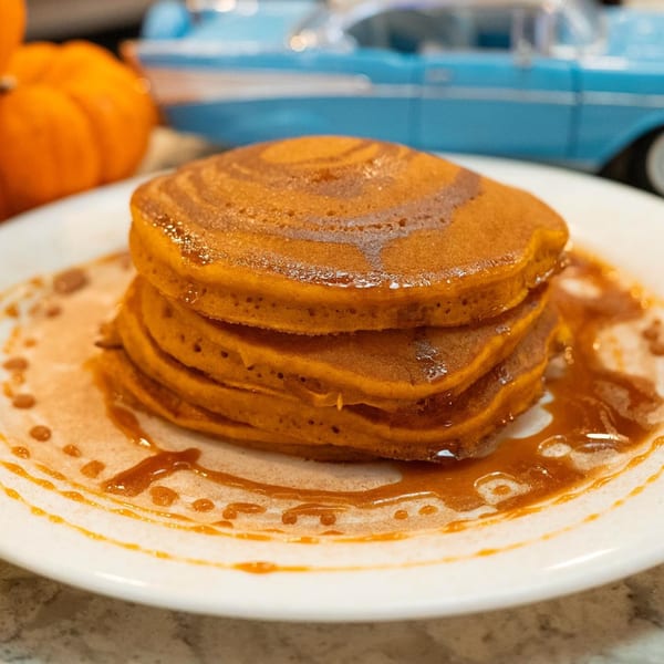 Stack of pumpkin pancakes drizzled with syrup on a white plate, with a toy car and pumpkin in the background.