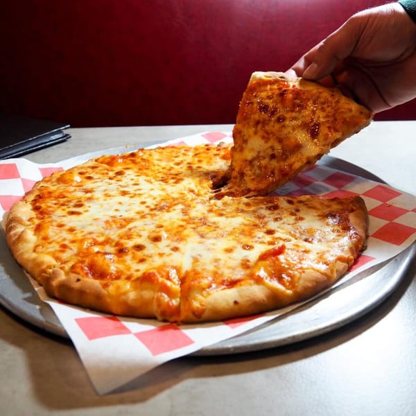 A hand lifts a cheesy slice from a whole pizza on a metal tray with checkered paper.