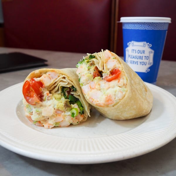 Two shrimp salad wraps with tomatoes and greens on a white plate, next to a blue diner cup.